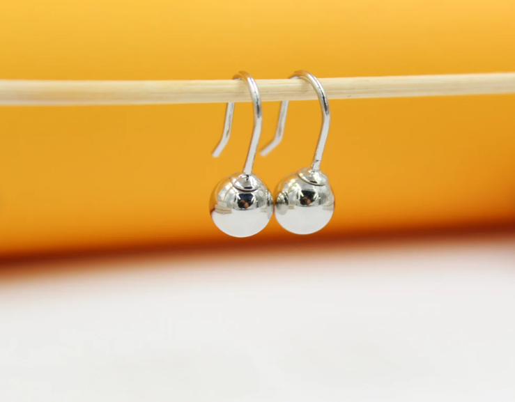 A pair of polished silver ball drop earrings hangs delicately from a wooden stick against a vibrant yellow and white background.