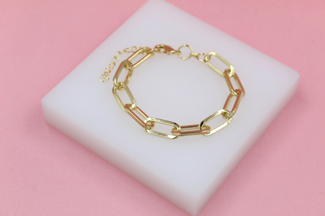A polished gold paperclip chain bracelet rests elegantly on a white display block against a soft pink background.