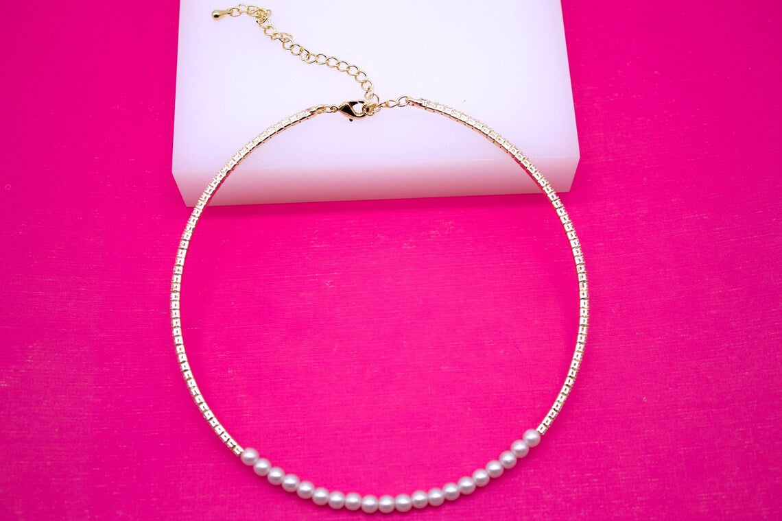 A gold chain necklace featuring sparkling crystals and a delicate line of white pearls rests on a vibrant pink background.