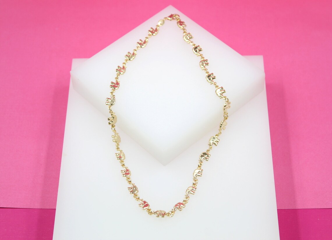 A gold elephant charm necklace is displayed on a white display block against a vibrant pink background.