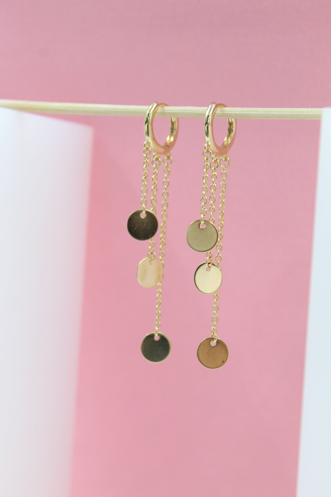 A pair of elegant gold hoop earrings featuring delicate dangling chains with small coin charms hang against a soft pink and white backdrop.