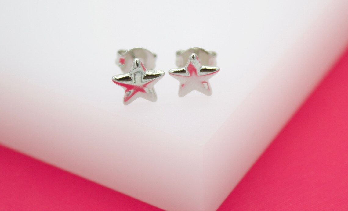 A pair of polished silver star stud earrings rests against a white and pink display.