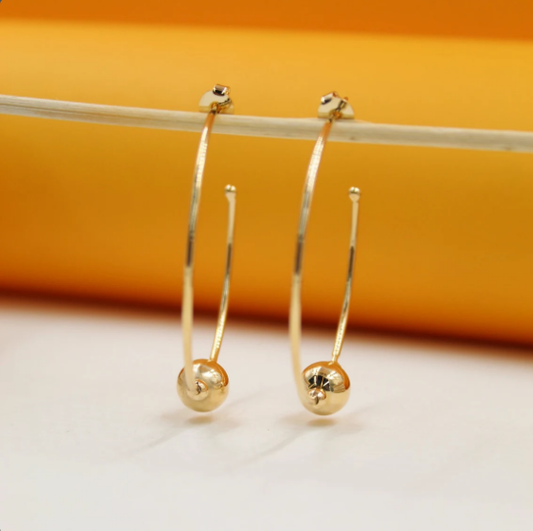 A pair of elegant gold hoop earrings with a shiny spherical charm hangs delicately from a thin stick against a vibrant orange and white background.