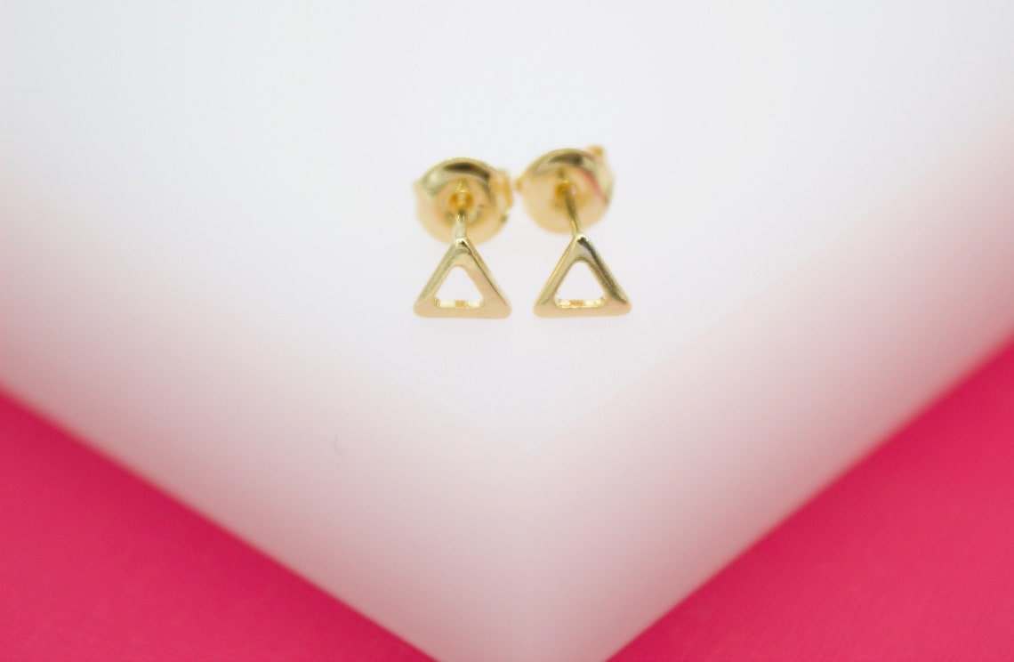 A pair of minimalist gold triangle stud earrings rests neatly on a white display block against a vibrant pink background.