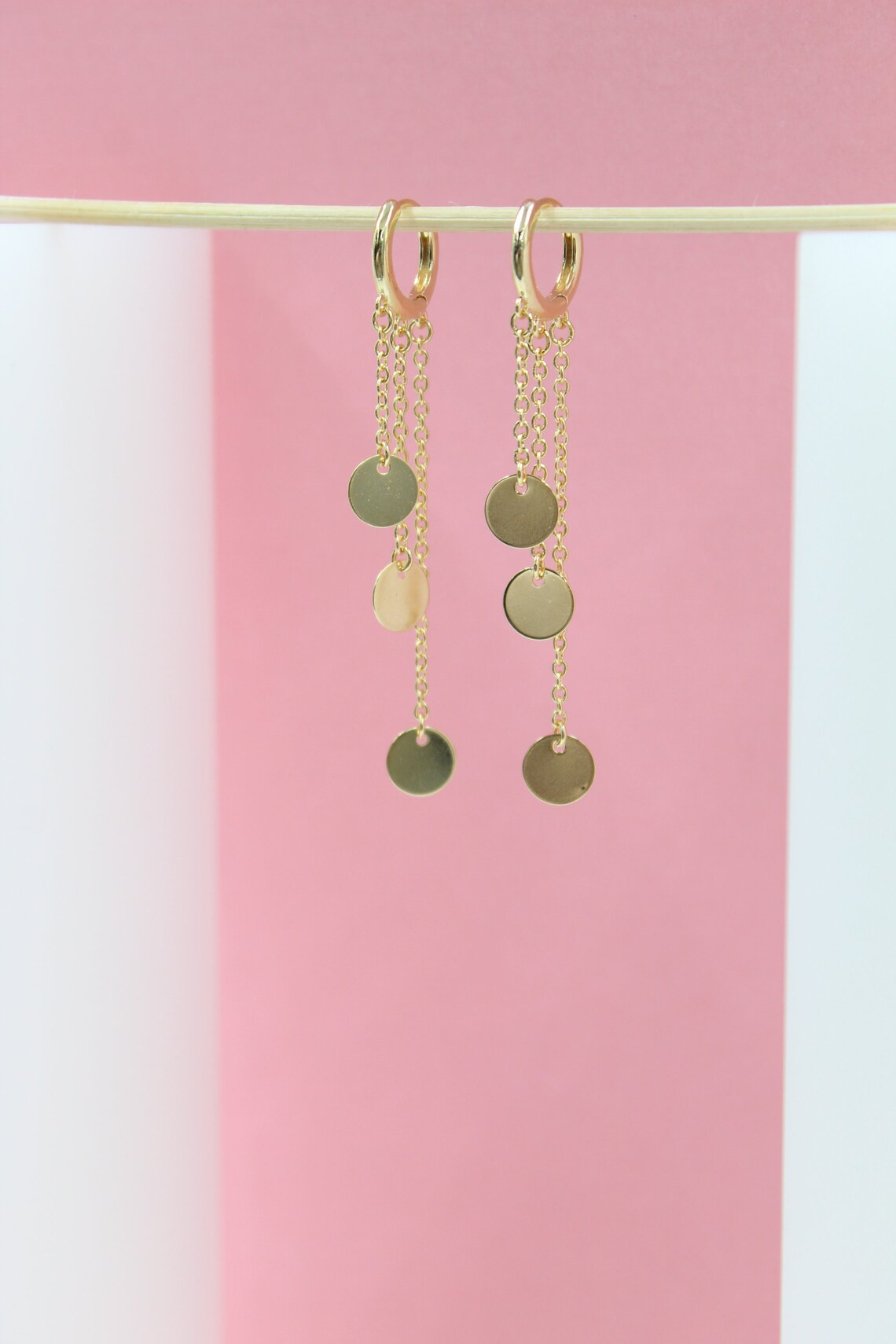 A pair of elegant gold chain dangle earrings with small circular charms hangs gracefully against a minimalist pink and white backdrop.
