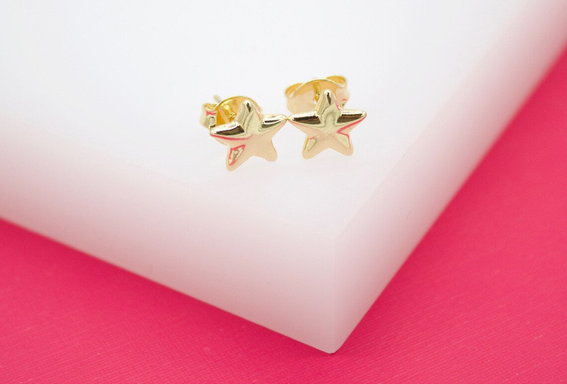 Two shiny gold star-shaped stud earrings rest on a white display stand against a vibrant pink background.