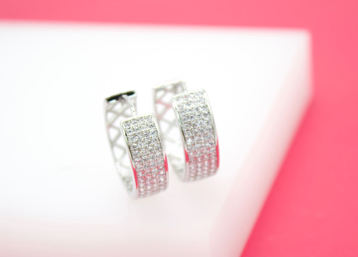 A pair of silver hoop earrings encrusted with sparkling cubic zirconia stones rests on a white display surface against a vibrant pink background.