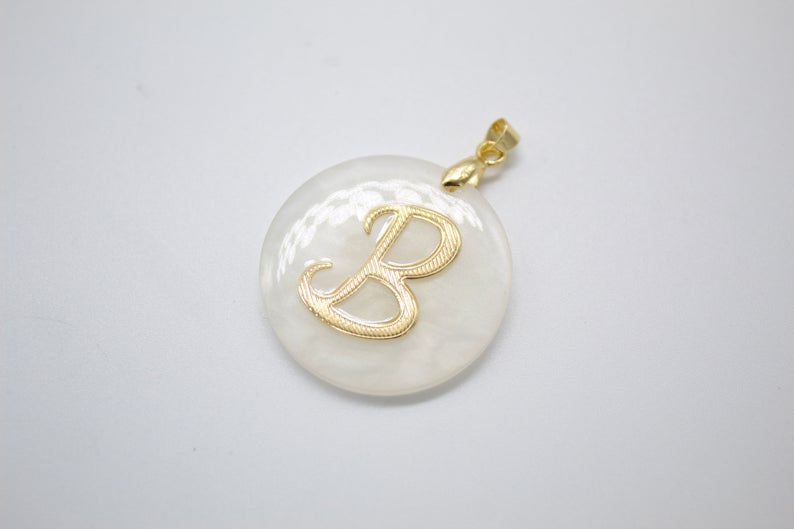 A round mother-of-pearl pendant features a gold initial B and rests against a plain white background.