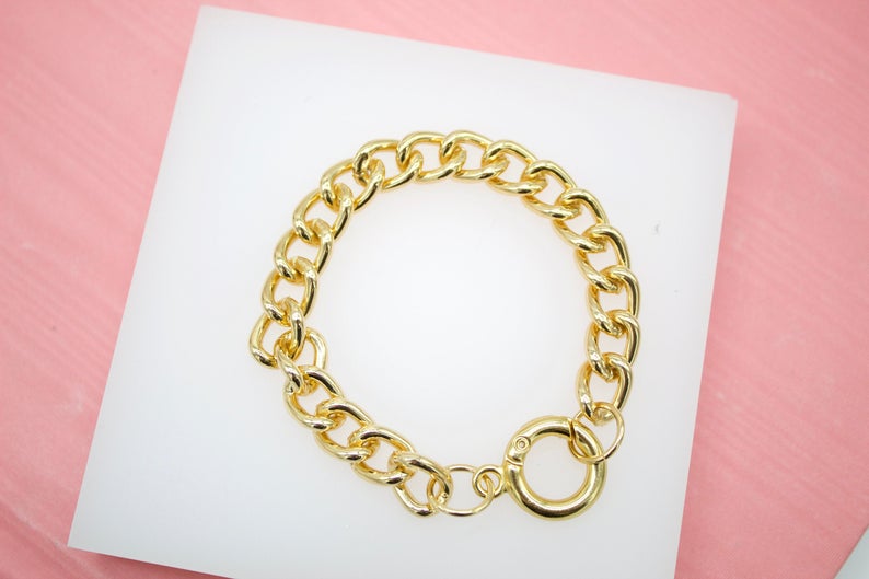 A thick gold chain bracelet rests on a white display surface against a pink background.