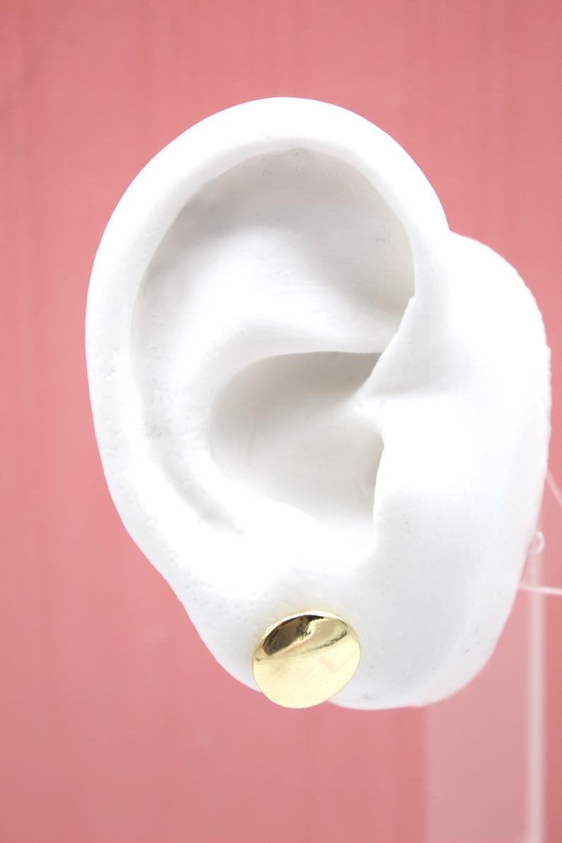 A white mannequin ear displays a shiny gold disc stud earring against a solid pink background.