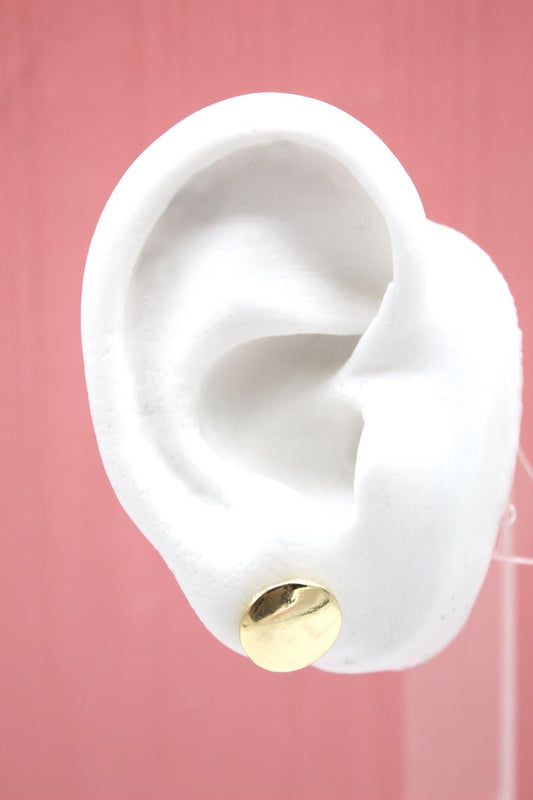 A white mannequin ear displays a shiny gold disc stud earring against a solid pink background.