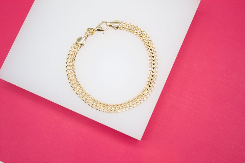 A delicate gold chain bracelet rests on a white display surface against a vibrant pink background.