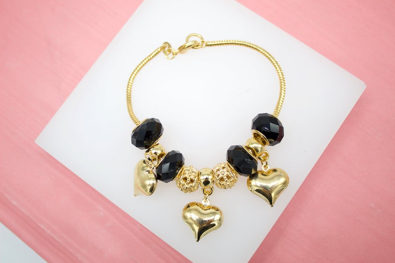 A gold snake chain charm bracelet displays black crystal beads and dangling gold heart pendants on a white display surface.