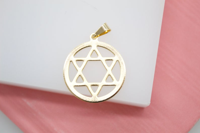 A shiny gold Star of David pendant rests on a white display surface against a pink background.
