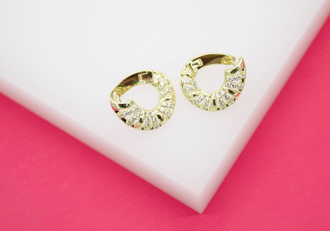 A pair of gold huggie hoop earrings featuring sparkling pave-set stones rest on a white display block against a vibrant pink background.