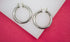 A pair of polished silver hoop earrings rests on a white display against a vibrant pink background.