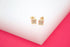 A pair of gold stud earrings featuring a square design with embedded stones rests on a white display surface against a vibrant pink background.