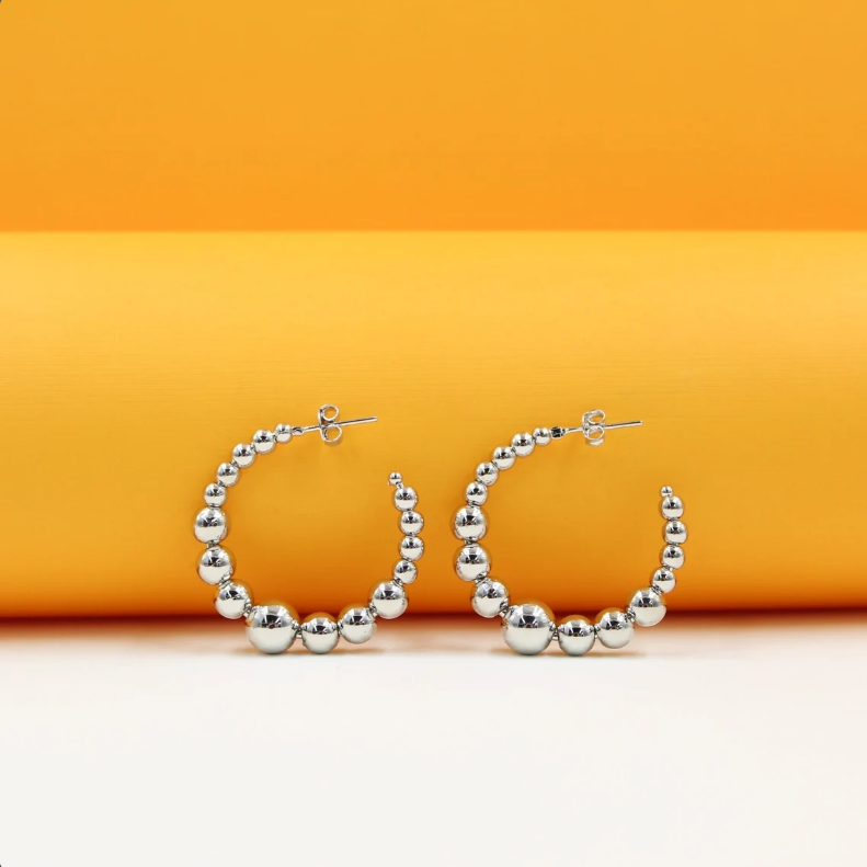 A pair of silver ball hoop earrings rests on a white surface against a bright orange background.