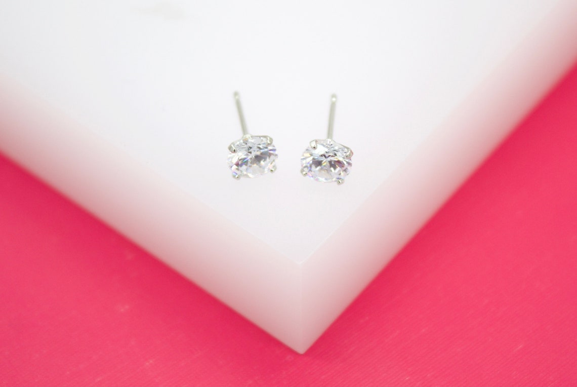 Two sparkling solitaire diamond stud earrings rest on a white display surface against a vibrant pink background.