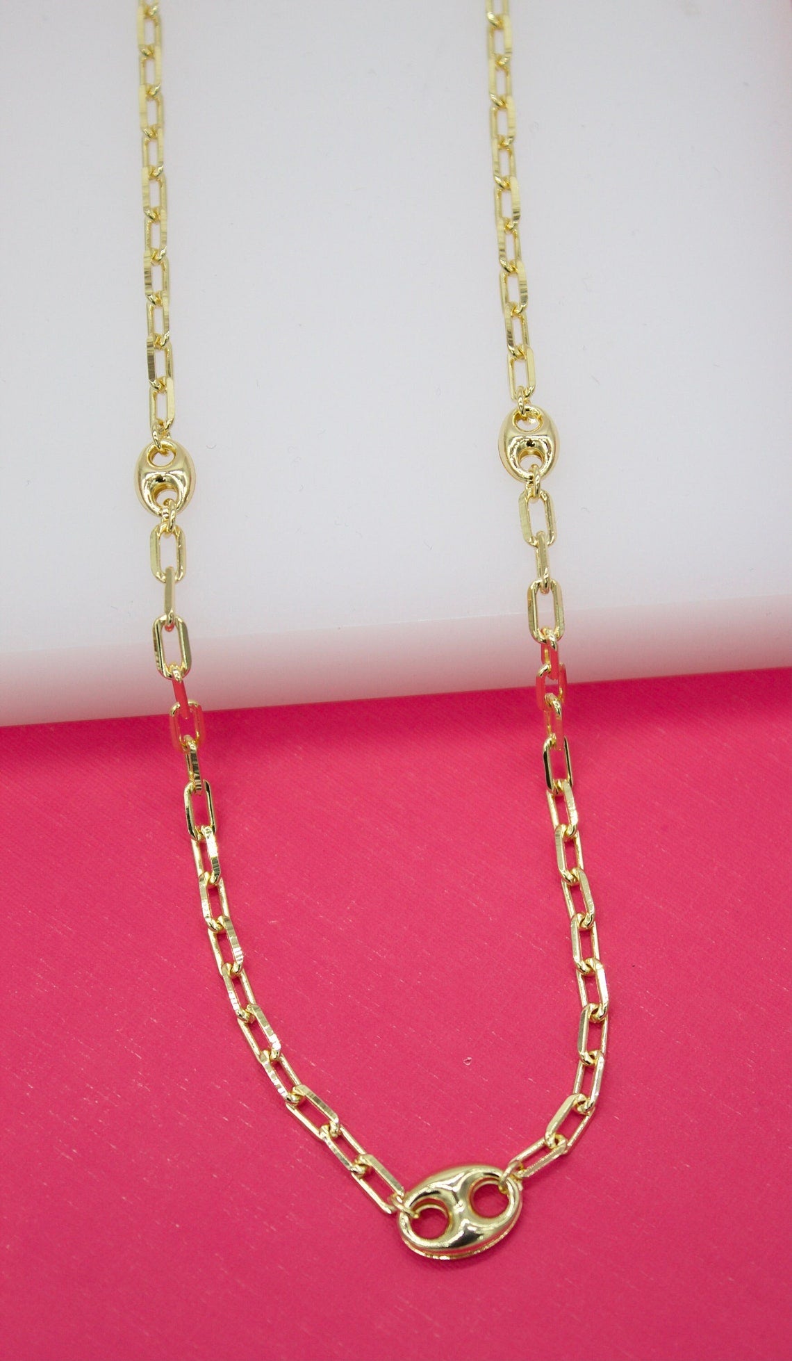 A shiny gold chain necklace with interspersed oval links drapes against a vibrant pink and white background.