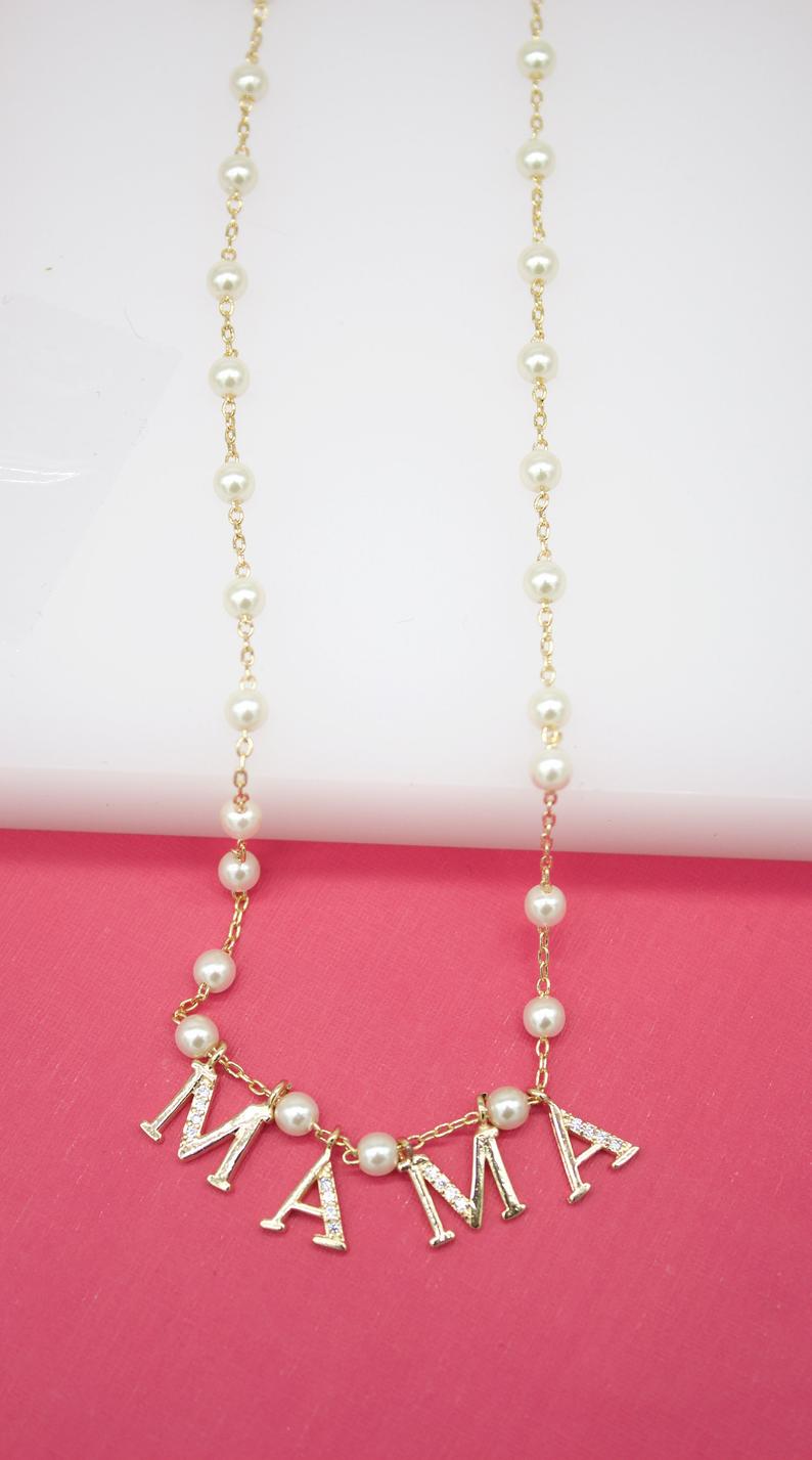 A delicate gold pearl necklace features rhinestone-encrusted letters spelling out "MAMA" against a pink background.