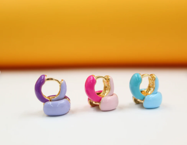 Three pairs of colorful enamel huggie hoop earrings in purple, pink, and blue rest on a white surface against a warm yellow background.