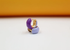 A pair of gold huggie hoop earrings featuring purple and light lavender enamel rests on a white surface against an orange backdrop.
