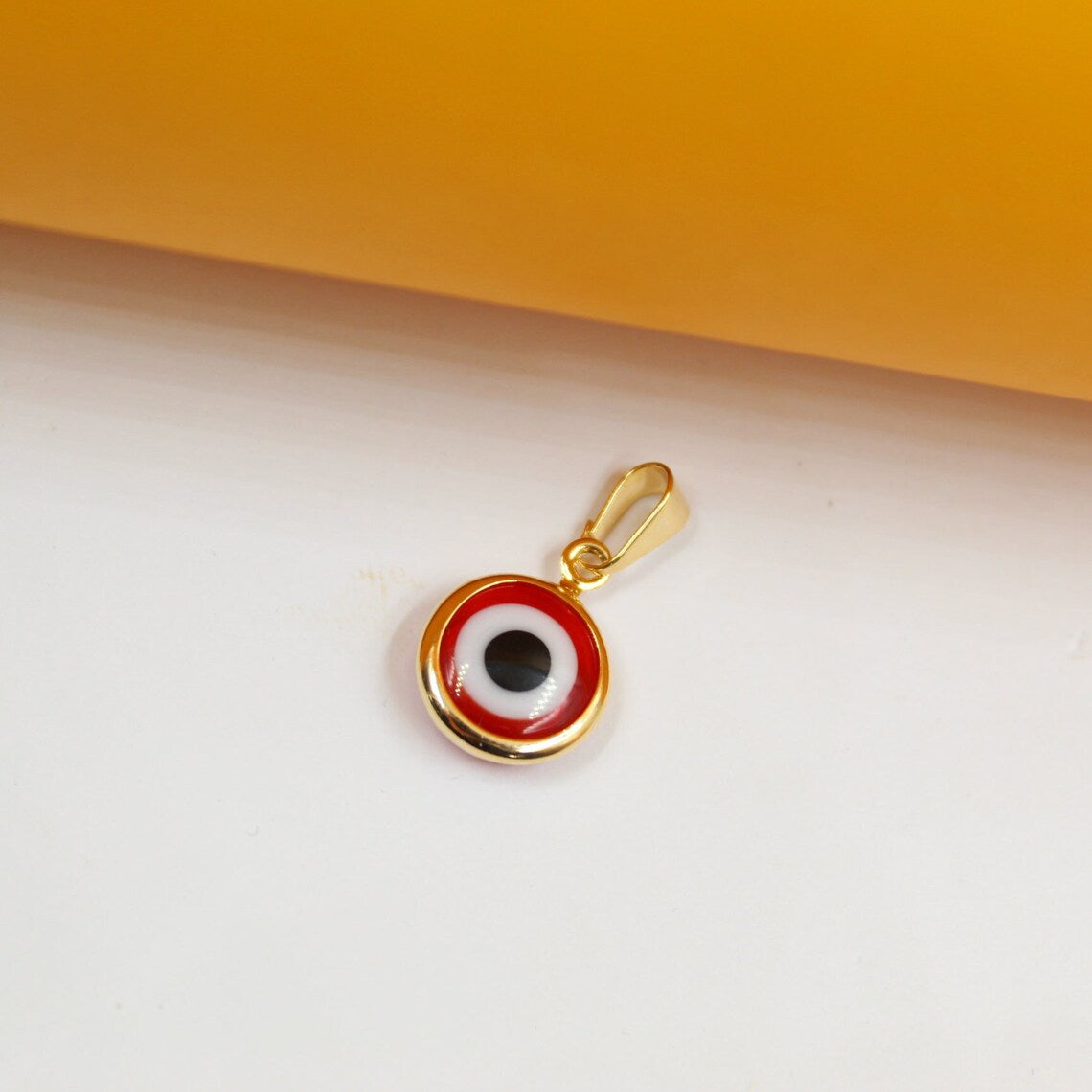 A gold Greek evil eye pendant rests on a clean white surface against a warm yellow backdrop.