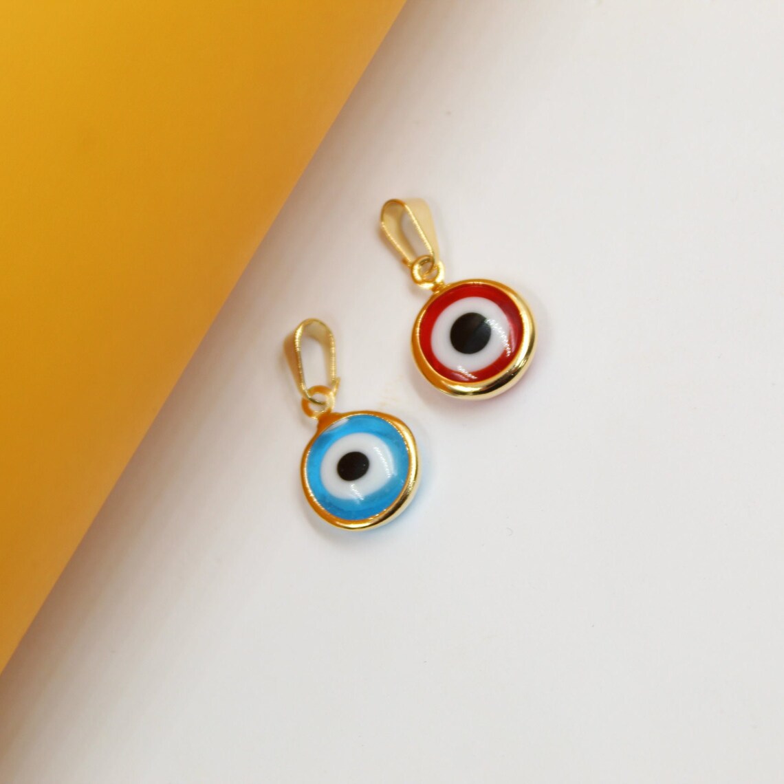 Two gold evil eye pendants rest on a white surface next to a yellow background, showcasing a vibrant blue charm and a striking red charm.