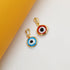 Two gold evil eye pendants rest on a white surface next to a yellow background, showcasing a vibrant blue charm and a striking red charm.