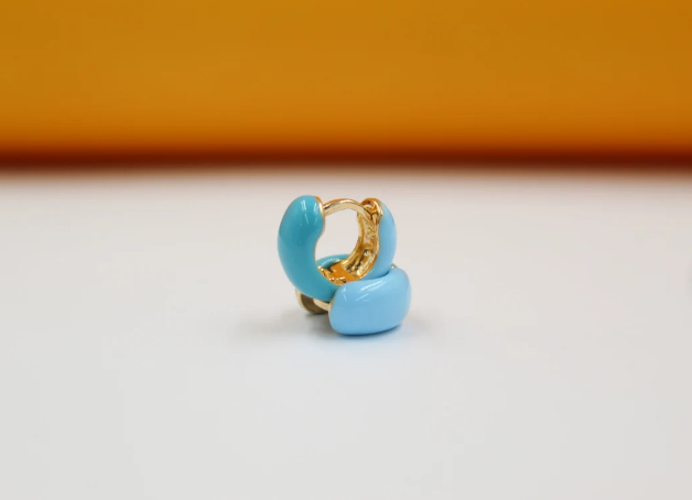 A pair of vibrant light blue enameled gold hoop earrings sits prominently on a white surface against an orange backdrop.