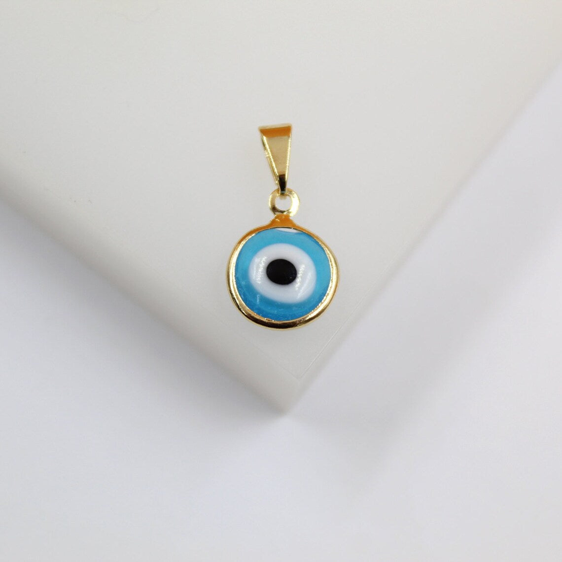 A gold evil eye pendant rests on a clean white surface, featuring vibrant blue and white enamel circles with a central black pupil.