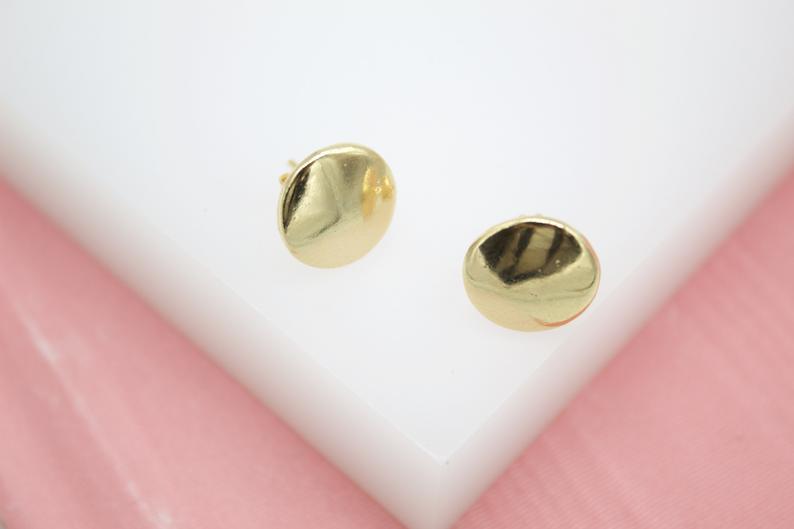 A pair of shiny gold-plated stud earrings rests on a white display block against a soft pink background.