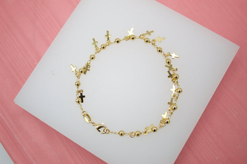A delicate gold chain bracelet features alternating small beads and hanging cross charms, displayed on a white surface.