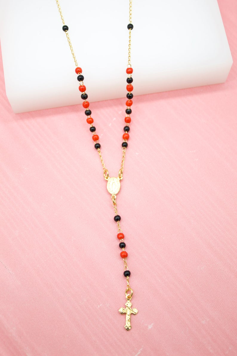 A gold rosary necklace featuring alternating red and black beads rests against a pink and white background.