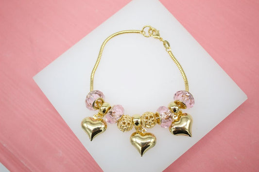A gold charm bracelet featuring pink crystal beads and dangling heart charms rests on a white display surface.