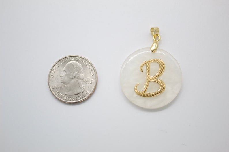 A round white pendant with a gold initial B rests beside a US quarter for scale.