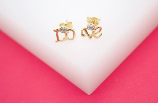 A pair of gold love stud earrings featuring sparkling cubic zirconia stones rests on a white and pink display.