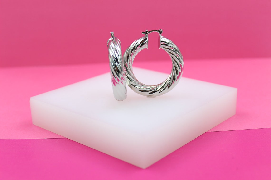 A pair of chunky silver twist hoop earrings rest on a white display block against a vibrant pink backdrop.