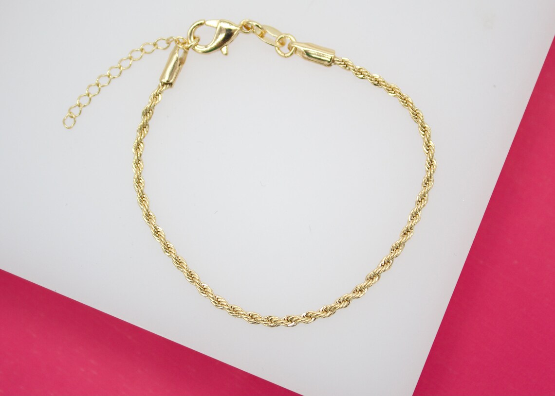 A thin gold rope chain bracelet lies on a white surface against a pink background, showcasing its lobster clasp and adjustable extension chain.
