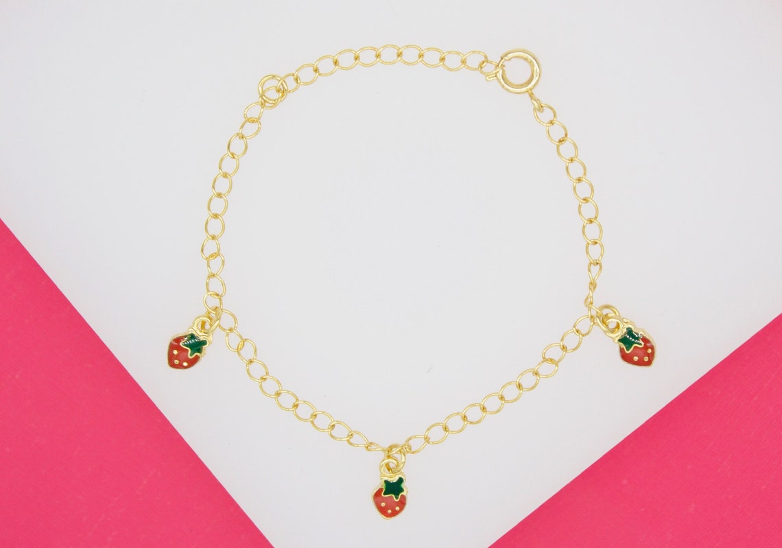 A gold chain bracelet features three hanging enamel strawberry charms against a pink and white background.