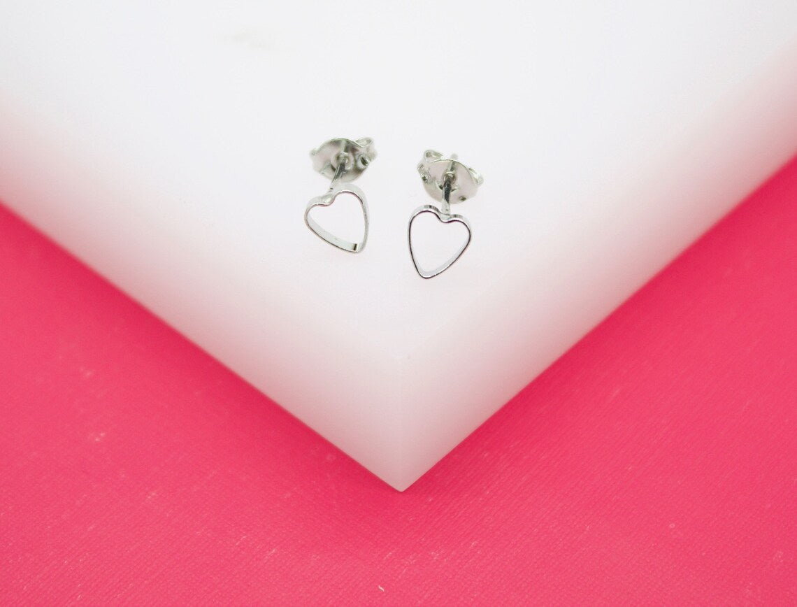 A pair of delicate silver heart-shaped stud earrings rests on a white display stand against a vibrant pink background.