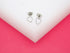A pair of delicate silver heart-shaped stud earrings rests on a white display stand against a vibrant pink background.