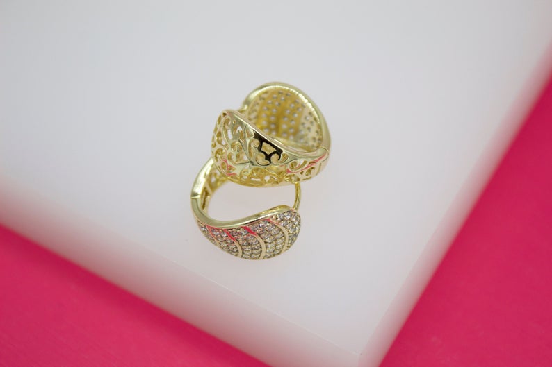 A pair of gold huggie hoop earrings featuring intricate filigree work and sparkling pavé stones rests on a white and pink surface.