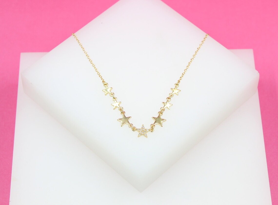 A delicate gold star pendant necklace displays sparkling pavé accents on a white display block against a vibrant pink background.