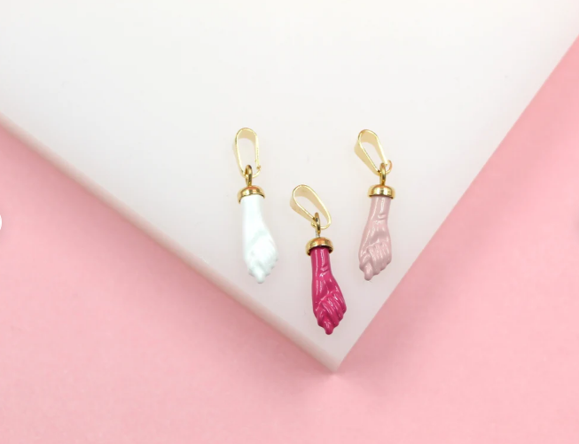 Three colorful figa charm pendants in white, fuchsia, and pink enamel with gold bails rest on a white display against a pink background.