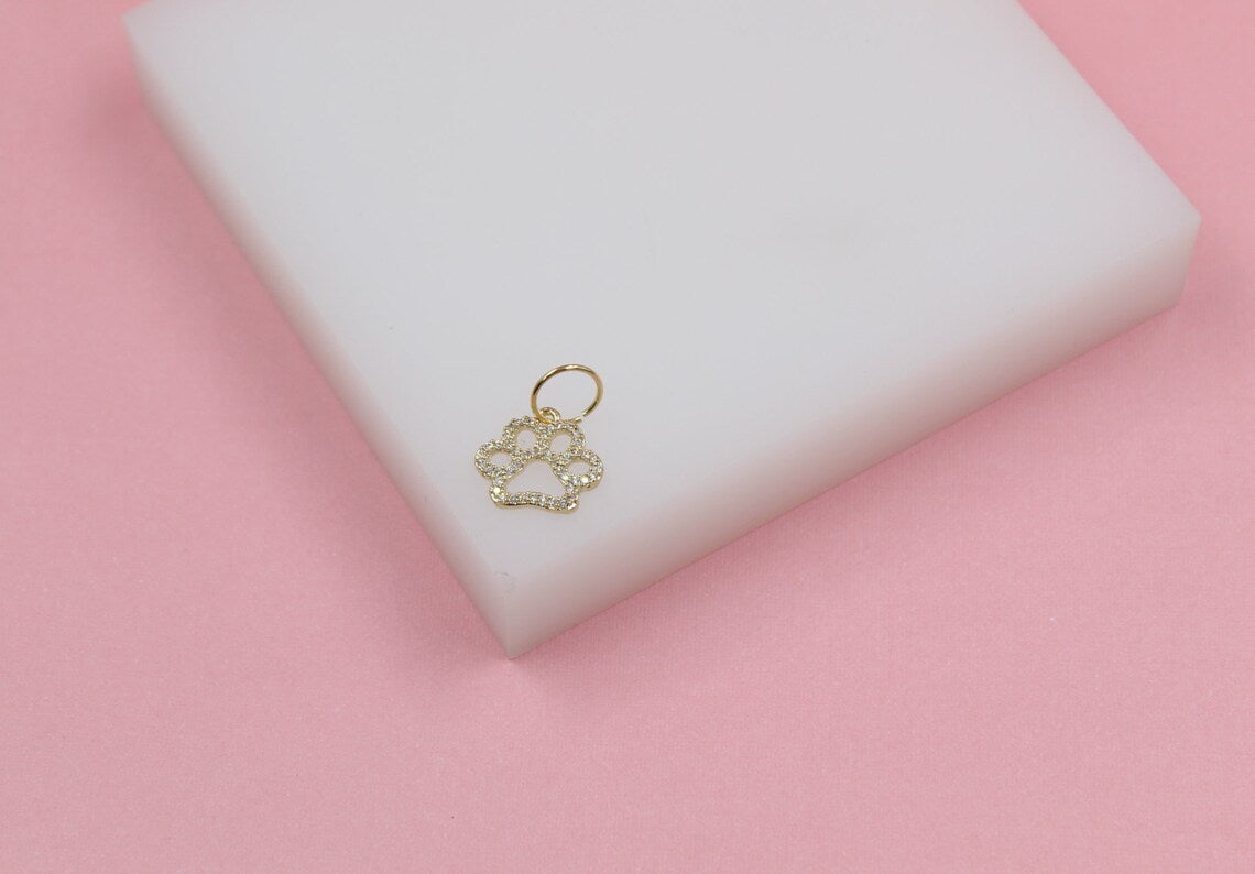 A gold paw print charm encrusted with small clear gemstones rests on a white block against a pink background.