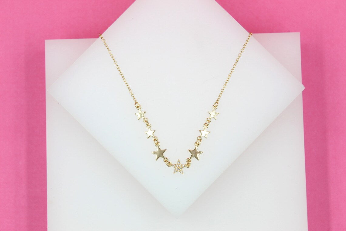 A delicate gold star pendant necklace rests on a white display stand against a vibrant pink background.