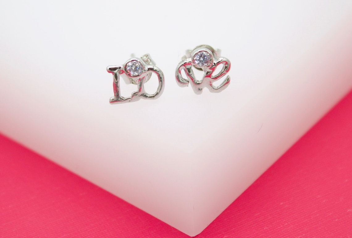 A pair of sterling silver 'LOVE' stud earrings sparkle with embedded cubic zirconia stones on a white and pink display.