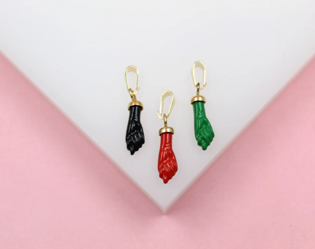 Three enamel figa hand charms in black, red, and green with gold bails rest on a white display against a pink background.